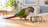 Green-cheeked conure beside a food bowl with a few scattered food pieces on a light wooden surface.